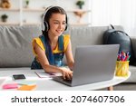 Small photo of Portrait of little girl in wireless headset using laptop, studying online at home, interested happy student typing on keyboard looking at pc screen, watching webinar, online course, doing homework