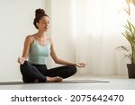 Small photo of Morning Meditation. Beautiful Calm Woman Meditating At Home In Lotus Position, Peaceful Young Brunette Lady Practicing Yoga, Sitting With Closed Eyes On Fitness Mat In Light Room, Copy Space