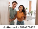 Portrait of excited cheerful couple inviting guest friends to enter their home, happy young guy and lady standing in doorway of contemporary flat, millennial family holding door looking out together
