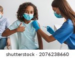 Covid-19 Vaccination. Black Girl Getting Vaccinated Gesturing Thumbs Up Approving Vaccine, While Female Medical Worker Injecting Her Arm With Syringe Sitting In Clinic Indoors, Wearing Face Mask