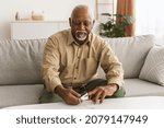 Sick Mature African Male Using Pulse Oximeter Measuring Saturation Level With Medical Device Siting On Sofa At Home. Pulseoxymetry, Coronavirus Disease In Older Age Concept