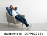 Calm young Arab man relaxing in armchair against white studio wall, copy space. Handsome middle Eastern guy enjoying peaceful weekend morning, resting with hands behind head