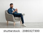 Joyful young Arab guy sitting in armchair with smartphone, working remotely or video chatting against white studio wall, empty space. Middle Eastern man watching movie, having online meeting