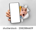 Wow, Unbelievable Offer. Shocked young guy holding smartphone with white empty cell screen, showing device close to camera breaking through paper sheet. Gadget display with copy space, mockup template
