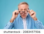 Poor Eyesight. Senior Man Can't See, Squinting Eyes Wearing Glasses Having Problems With Vision, Looking At Camera At Blue Studio. Ophtalmic Issue, Bad Sight In Older Age, Macular Degeneration Concept