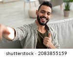 Small photo of Happy young Arab guy taking selfie and showing thumb up gesture, smiling at mobile phone camera, sitting on sofa at home. Portrait of positive middle Eastern man making photo of himself indoors