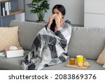 Small photo of Unhappy young indian lady sneezes in napkin, blows nose, suffers from fever, flu on couch in room interior with tea, honey and lemon. Home treatment for cough, runny, cold and covid-19, self-isolation