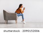Small photo of Side view of young Asian woman sitting in armchair, drinking hot aromatic coffee, relaxing against white studio wall, free space. Lovely millennial lady enjoying warm beverage on lazy day