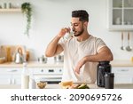 Small photo of Healthy nutrition for muscle gain and weight loss concept. Athletic young Arab man drinking protein shake or milk, standing near table with healthy wholesome products at kitchen, copy space