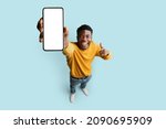 Cheerful african american young man in yellow showing modern cellphone with blank white screen and smiling on blue studio background, mockup, recommending newest mobile app, top view, copy space