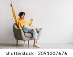 Joyful young Asian guy in wireless headphones sitting in armchair, having fun, listening to music, raising his arm, dancing to favorite song, enjoying cool soundtrack against white wall, empty space