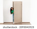 Portrait of cheerful young woman standing in doorway of modern apartment, millennial female homeowner holding slightly open ajar door looking out and smiling, greeting visitor, full body length