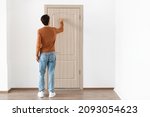 Small photo of Rear back view of young man knocking on the wooden closed door, male visitor standing in front entrance, guest wants to come in, free copy space, full body length