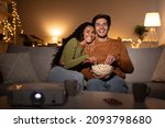 Couple Watches Movie Via Domestic Cinema Projector And Eats Popcorn From Bowl During Evening At Home, Sitting On Sofa In Living Room. Front View, Selective Focus On Happy Spouses