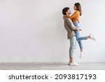 Side view of romantic young Asian guy holding and hugging his beloved girlfriend against white studio wall, copy space. Full length fo millennial spouses expressing love and affection