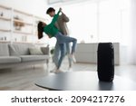 Small photo of Millennial Asian couple dancing to popular music at home, selective focus on portable wireless speaker on table, copy space. Young family moving to their favorite song, using modern device