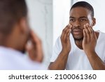 Small photo of Dull Skin. Worried Young Black Man Looking At Mirror In Bathroom And Touching Face, Concerned Millennial African American Guy Having Dark Circles Under Eyes, Unhappy With His Look, Selective Focus