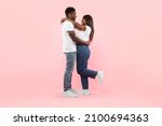 Couple In Love. Portrait of joyful black man hugging his girlfriend, standing together isolated over pink studio background. Casual guy and lady smiling, profile side view, full body length, banner