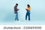 Small photo of Modern Technology And Relationship. Full body length of cheerful black girlfriend and boyfriend using their smartphones, texting each other, walking isolated over blue studio background, banner