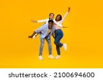 Spending Time With Family Is Fun. Full body length of excited African American man, woman and girl laughing and posing isolated on yellow studio wall. Cheerful father carrying daughter on back, banner