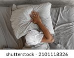 Closeup of irritated young black man lying in bed and covering head and ears with pillow, hearing and suffering from too loud sound. Angry millennial guy can't sleep, tired of noisy neighbors