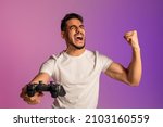 Small photo of Triumphant young Arab guy winning online esports competition, playing video game, shouting OMG, overjoyed about his gaming victory, making YES gesture in neon light