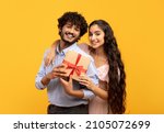 Small photo of Portrait of loving indian couple holding wrapped gift for Valentine's Day together, posing on yellow studio background. Young woman and her boyfriend with romantic holiday present