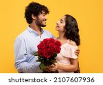 Small photo of Lovers holiday. Loving indian couple holding bouquet of flowers, embracing and looking at each other on yellow background. Young guy pampering his girlfriend with roses on Valentine's Day