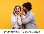 Small photo of Shock, gossip, share advice. Young indian man whispering to lady on ear, happy woman with open mouth excited and surprised, posing on yellow studio background