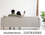 Back view of hugging black lovers sitting on couch at home, watching TV, unrecognizable african american man aiming remote controller towards copy space on white wall, panorama, full length