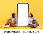Happy indian couple sitting near big smartphone with blank white screen, demonstrating copy space for app or ad design, posing over yellow background, mockup banner