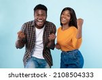 Small photo of Wow, Yes. Portrait of overjoyed young black man and woman cheering and shaking clenched fists, looking at camera. Happy couple celebrating win posing standing isolated over blue studio background wall