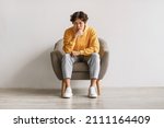 Negative emotions. Young Asian man sitting in armchair with thoughtful face expression, feeling sad or tired, having problem, suffering from depression or stress against white studio wall