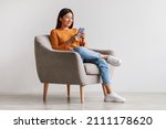 Small photo of Charming young Asian woman using cellphone, communicating on web, working or learning online, sitting in armchair against white wall, free space. Lovely millennial lady chatting on smartphone