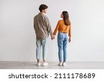 Full length of young Asian man and woman standing with their backs to camera, looking at each other and holding hands against white studio wall. Millennial couple expressing affection