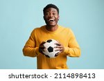 Happy millennial black guy in casual with soccer ball posing on blue studio background, emotional african american young man football fan going to game, copy space. Sport concept