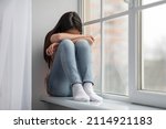 Small photo of Upset asian school-aged girl sitting on windowsill and crying, female kid feeling lonely or insulted, got quarrel with parents or friends, home interior, panorama with copy space
