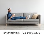 Small photo of Rest Concept. Happy Arab guy drinking coffee sitting on comfortable couch at home in living room. Cheerful casual man relaxing on sofa, enjoying weekend free time or break from work, full body length