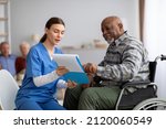 Small photo of Attractive young woman in blue workwear nurse helping senior black man in wheelchair with questionnaire, african american pensioneer filling papers at nursing home, having assistance