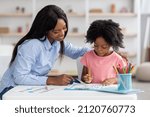 Small photo of Cute african american little girl with bushy hair exercising at daycare, sitting at table with female teacher and doing various tests, young woman psychologist working with child preschooler