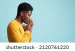 Small photo of Side view of praying african american young man in yellow, holding palms by his lips and whispering pray, asking for grace over blue studio background, panorama wuth copy space