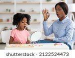 Pretty little black girl attending speech therapy session at clinic, looking at mirror, beautiful young african american woman speech-language pathologist working on sounds with kid, articulating