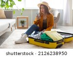 Small photo of Beautiful young woman staring at smartphone screen in terror, feeling shocked about cancelled flight, sitting near open suitcase with packed clothes at home. Travel ban during covid epidemic