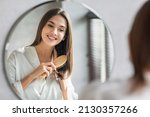 Small photo of Beauty Routine. Pretty Woman Combing Her Beautiful Hair With Brush While Standing Near Mirror In Bathroom, Attractive Young Lady Looking To Her Reflection And Smiling, Selective Focus With Free Space
