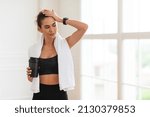 Small photo of After Training. Tired young woman with white towel on neck holding shaker with water swiping sweat from forehead resting after training routine standing indoors at gym or home