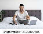 Portrait Of Young Arab Man Sitting In Bed With Laptop, Notepad And Coffee, Happy Millennial Middle Eastern Guy Using Computer In Bedroom For Online Work And, Enjoying Freelancing, Copy Space