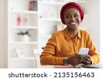 Positive beautiful young muslim black woman in red headscarf using brand new mobile phone, looking at copy space and smiling, using newest mobile application for business, home interior