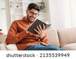 Portrait of young Middle Eastern man wearing eyeglasses holding paper book or diary, sitting on couch at home in living room, reading literature or checking his working schedule plan, free copy space