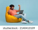 Happy curly bearded millennial hindu guy in casual gambling online, sitting at bean bag over blue studio background, using brand new laptop, raising hands up, celebrating success, copy space