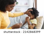 Seasonal Flu Concept. Worried Black Mom Taking Care Of Her Ill Child At Home, Caring Mother Touching Daughter's Forehead And Looking At Thermometer, Checking Kid's Temperature, Closeup Shot